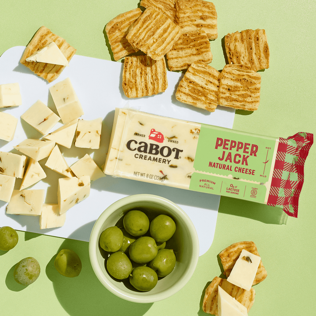 A block of Cabot Creamery Pepper Jack Cheese, cut cheese cubes, green grapes in a bowl, and rectangular crackers are arranged on a green background.