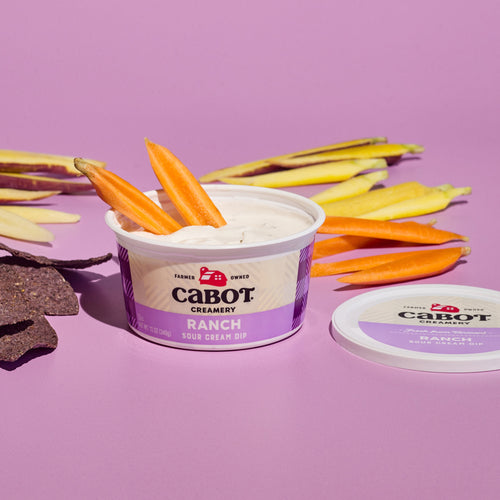 An open container of Cabot Creamery Ranch Dip with carrot sticks nestled inside sits against a pink background. The lid lies beside it, surrounded invitingly by multicolored carrot sticks and dark chips.