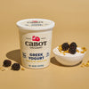 A tub of Cabot Creamery Reduced Fat Vanilla Bean Greek Yogurt is next to a bowl filled with yogurt, topped with blackberries and honey-glazed oats. The beige backdrop enhances the protein-rich treats packaging and toppings.