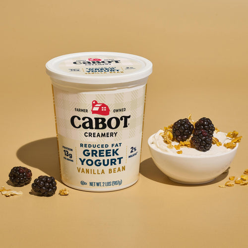 A tub of Cabot Creamery Reduced Fat Vanilla Bean Greek Yogurt sits next to a small bowl of yogurt topped with blackberries and granola, all on a beige background—an inviting scene featuring this high-protein snack.