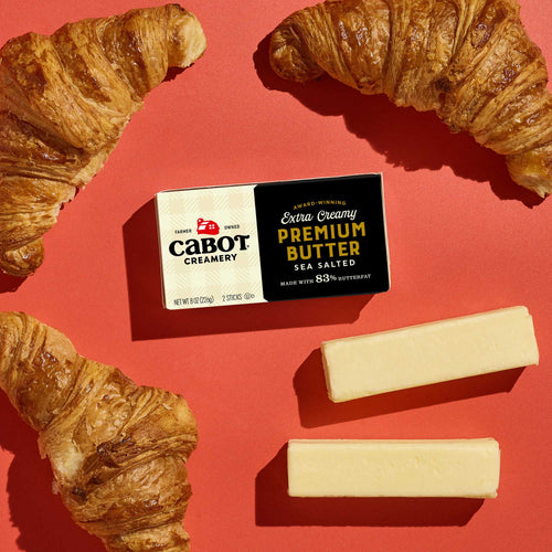 A package of Cabot Creamery Extra Creamy Sea Salted Butter and two butter sticks are displayed on a red surface, surrounded by three croissants.
