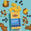 A block of Cabot Creamery Seriously Sharp Yellow Cheddar Cheese is shown with sliced pieces, surrounded by crackers, assorted nuts, and blueberries on a light blue background.