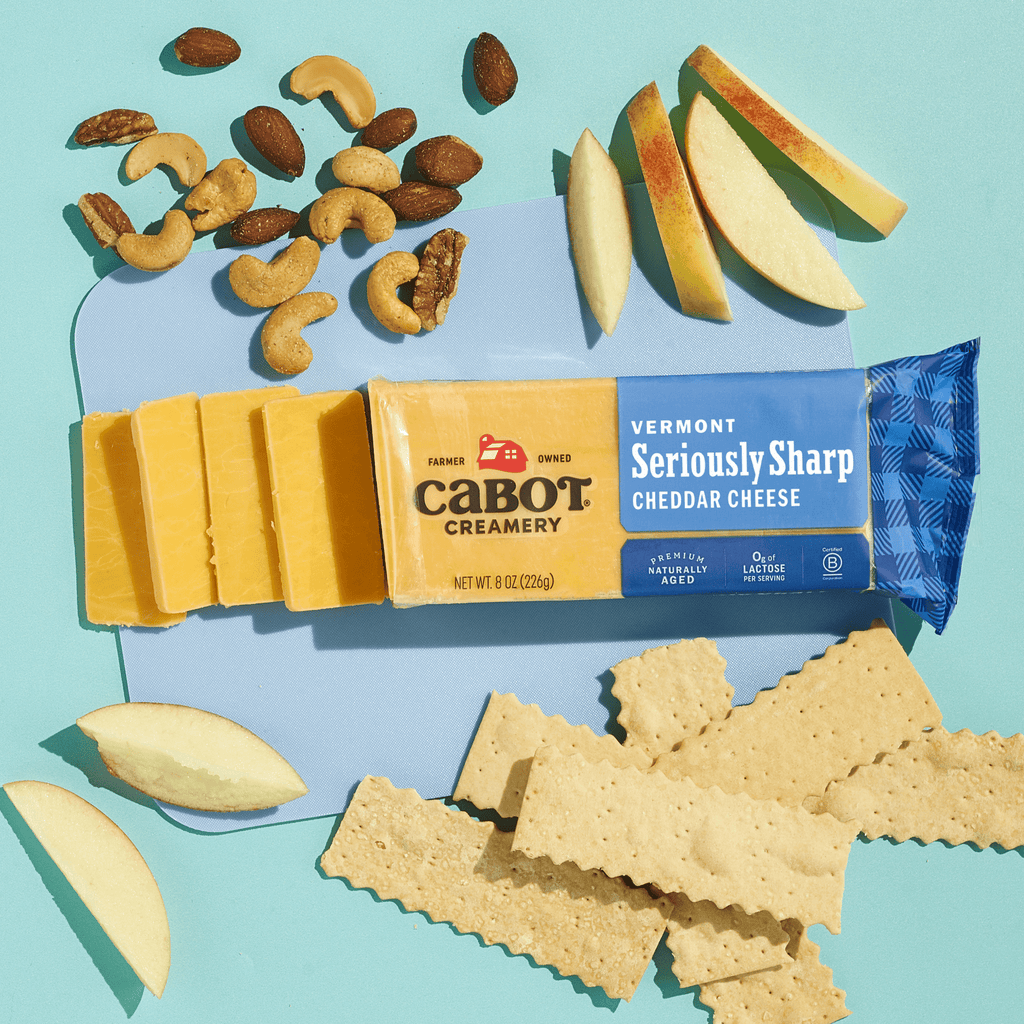 Cabot Creamery Seriously Sharp Yellow Cheddar Cheese, partly sliced, rests on a light blue surface with apple slices, assorted nuts, and rectangular crackers—a bold, award-winning flavor ideal for gluten-free snacking.