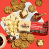 A wedge of Cabot Creamery Seriously Sharp Cheddar Cheese sits on a white board with cut pieces, surrounded by multigrain crackers, a honey jar with dipper, and a bowl of dark olives on a red background.