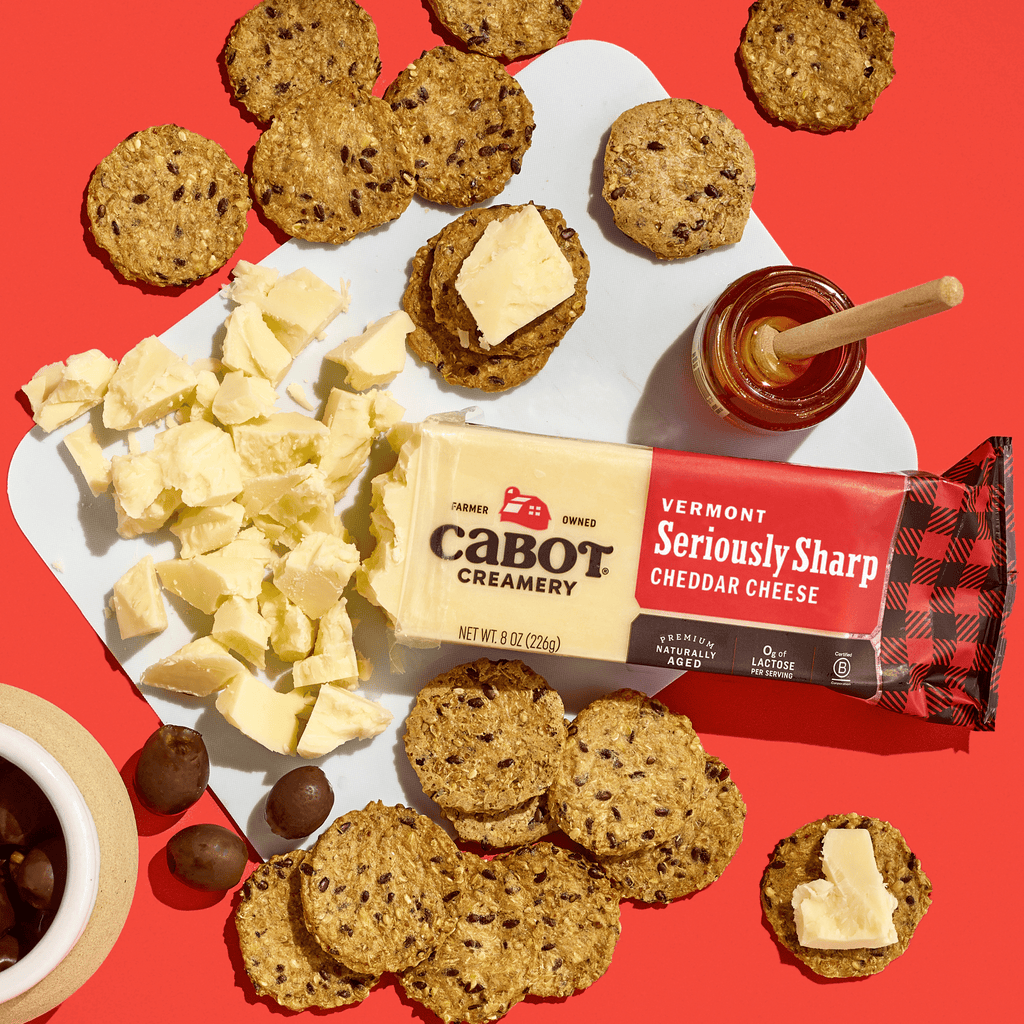 A wedge of Cabot Creamery Seriously Sharp Cheddar Cheese sits on a white board with cut pieces, surrounded by multigrain crackers, a honey jar with dipper, and a bowl of dark olives on a red background.