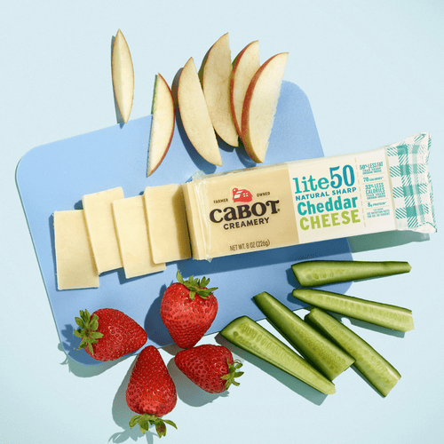 Cabot Creamery Lite50 Sharp Cheddar Cheese is sliced on a blue surface with apple slices, strawberries, and cucumber sticks for a tasty, low-fat snack.