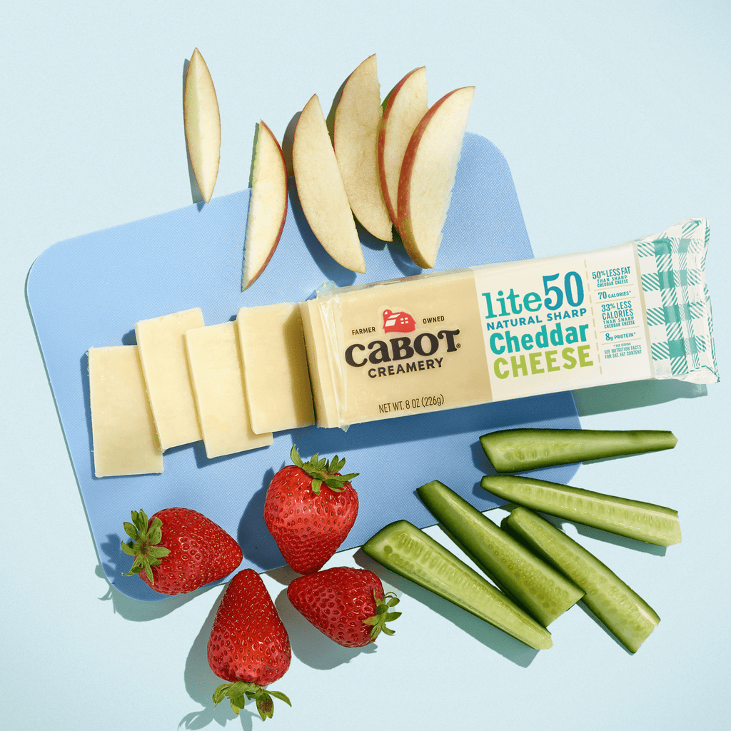 Cabot Creamery Lite50 Sharp Cheddar Cheese is sliced on a blue surface with apple slices, strawberries, and cucumber sticks for a tasty, low-fat snack.