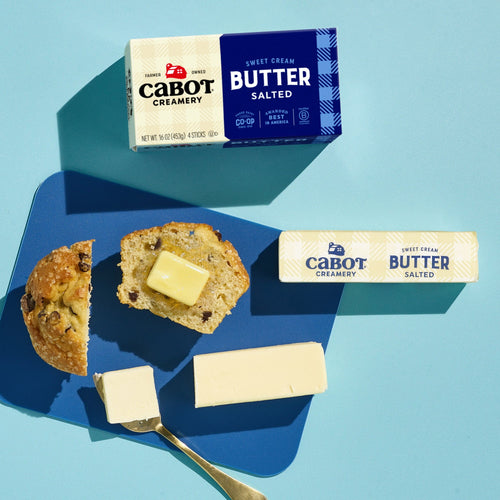 A blue surface with a muffin topped with Cabot Creamery Salted Butter on a blue plate, a stick of Cabot Creamery Salted Butter, a pat of the award-winning butter with a knife, and two butter packages labeled Cabot Creamery.