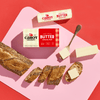 A loaf of seeded bread, several slices, and a stick of Cabot Creamery Unsalted Butter with a butter knife are arranged on a pink and red background.