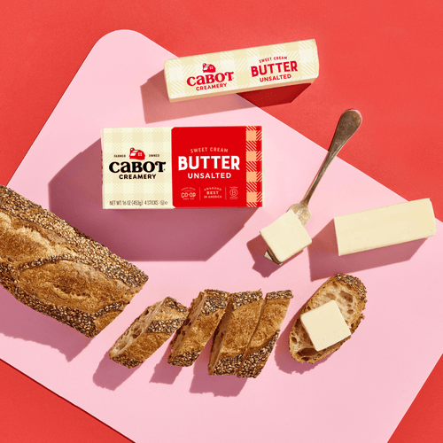 A loaf of seeded bread, several slices, and a stick of Cabot Creamery Unsalted Butter with a butter knife are arranged on a pink and red background.