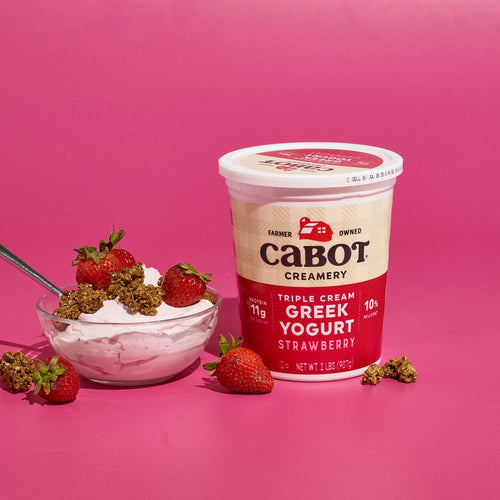 A container of Cabot Creamery Triple Cream Strawberry Greek Yogurt sits beside a bowl topped with strawberries and granola, plus a spoon. Fresh berries and granola pieces are scattered on a pink background. Savor this high protein treat!.