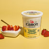 Cabot Creamery Triple Cream Vanilla Bean Greek Yogurt container next to a bowl of rich yogurt garnished with sliced strawberries and a spoon, all arranged on a yellow background with whole strawberries scattered nearby.