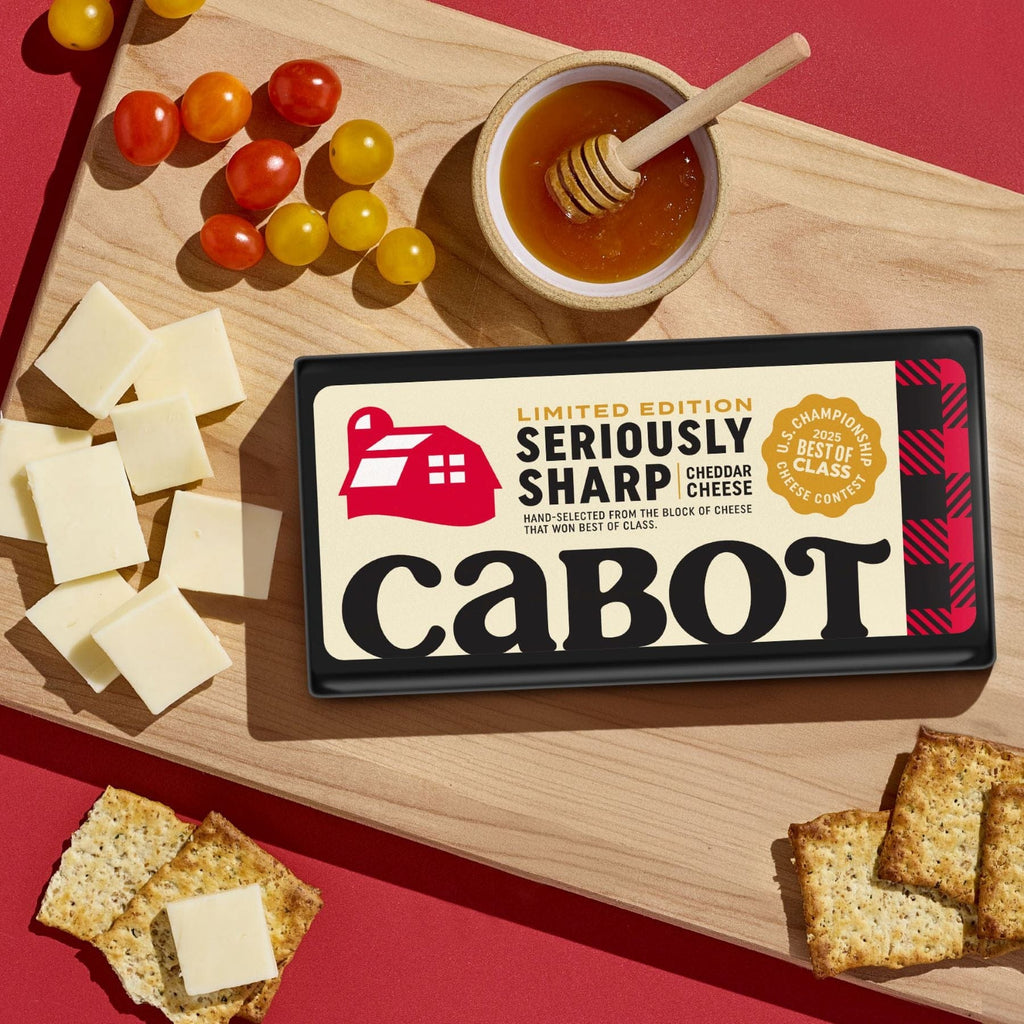 A block of Cabot Creamery Limited Edition Seriously Sharp Cheddar Cheese sits on a wooden board with cheese slices, cherry tomatoes, crackers, and a bowl of honey with dipper, all set against a red background.