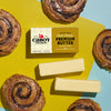Two sticks of Cabot Creamery Extra Creamy Unsalted Butter, with 83% butterfat for perfect baking, are placed beside cinnamon rolls on a yellow and teal background.