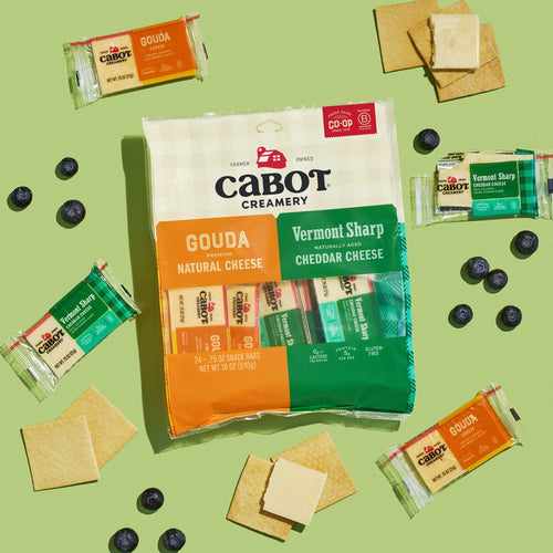 A package of Cabot Creamery Gouda & Sharp Variety Pack Cheese Snacks, with individual Gouda and Vermont Sharp Cheddar bars, is paired with crackers and blueberries on a light green background.