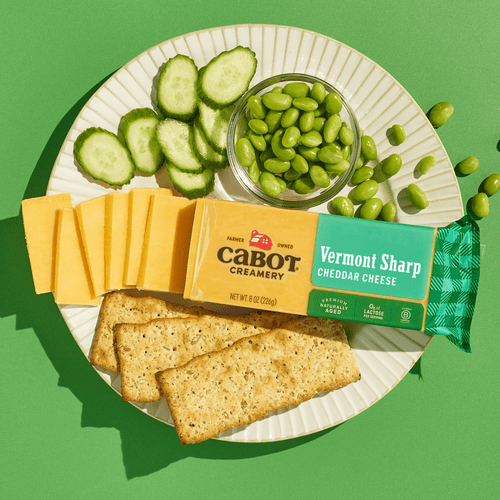 A plate with sliced Cabot Creamery Vermont Sharp Yellow Cheddar Cheese, cucumber rounds, edamame in a glass bowl, and rectangular crackers sits on a green background. The cheese packaging reads Cabot Creamery Vermont Sharp Yellow Cheddar Cheese.