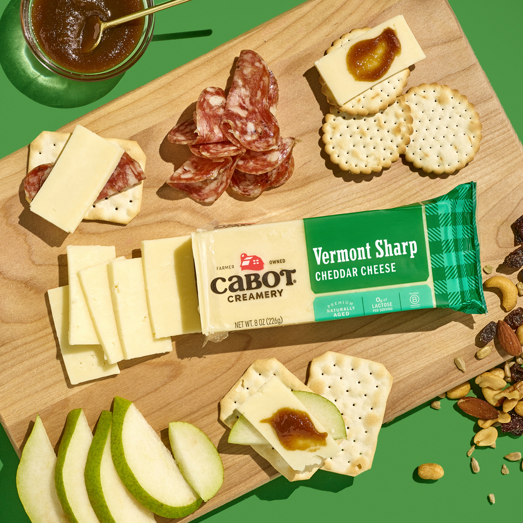 A wooden board with Cabot Creamery Vermont Sharp Cheddar Cheese, sliced cheese, round crackers, salami, green apple slices, mixed nuts, and a jar of fig spread on a bright green background.