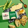 Cabot Creamery Vermont Sharp Cheddar Cheese cracker cuts are served on a black and white platter with cucumber sticks, crusty bread, and jam. The cheese’s packaging is visible on a green background—ideal for snacking or cooking.