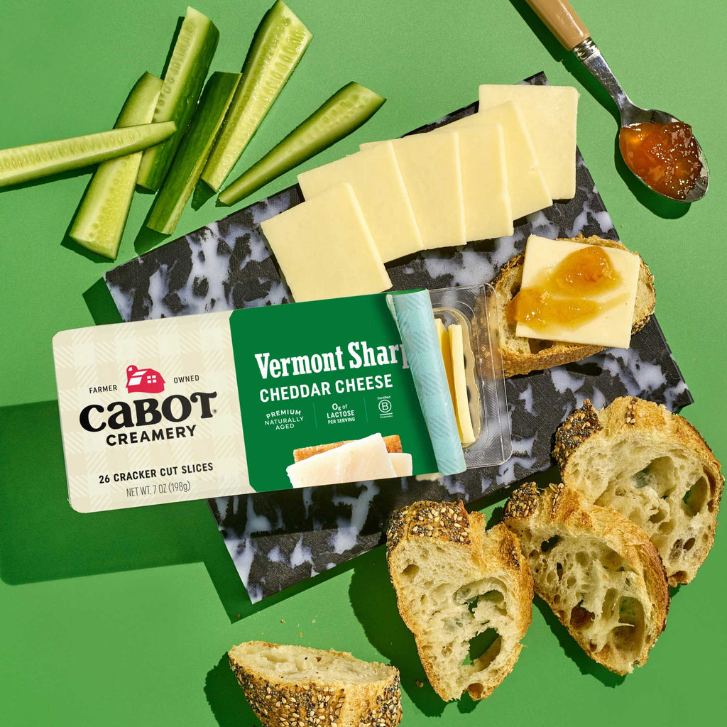 Cabot Creamery Vermont Sharp Cheddar Cheese cracker cuts are served on a black and white platter with cucumber sticks, crusty bread, and jam. The cheese’s packaging is visible on a green background—ideal for snacking or cooking.