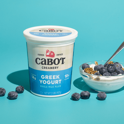 A container of Cabot Creamery Plain Greek Yogurt sits beside a bowl of yogurt topped with blueberries and granola on a blue surface, scattered with blueberries. A spoon rests in the bowl, ready to inspire your next recipe.