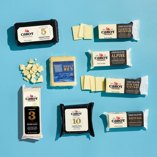 Cabot Creamery’s Cheese, Please Cheddar Medley features assorted cheddar blocks and slices arranged on a light blue surface, with several varieties unwrapped and crumbled aged cheddar pieces visible on the left.