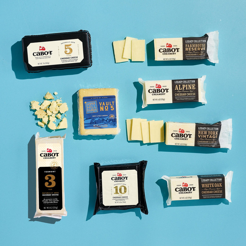 Cabot Creamery’s Cheese, Please Cheddar Medley features assorted cheddar blocks and slices arranged on a light blue surface, with several varieties unwrapped and crumbled aged cheddar pieces visible on the left.