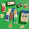 An overhead view of Cabot Creamery’s Classic Vermont Charcuterie Gift Box with cheddar, crackers, salami, jam jars, and Stowe Mercantile Kitchen Simply Salted Crackers on a green background—perfect for a charcuterie basket.