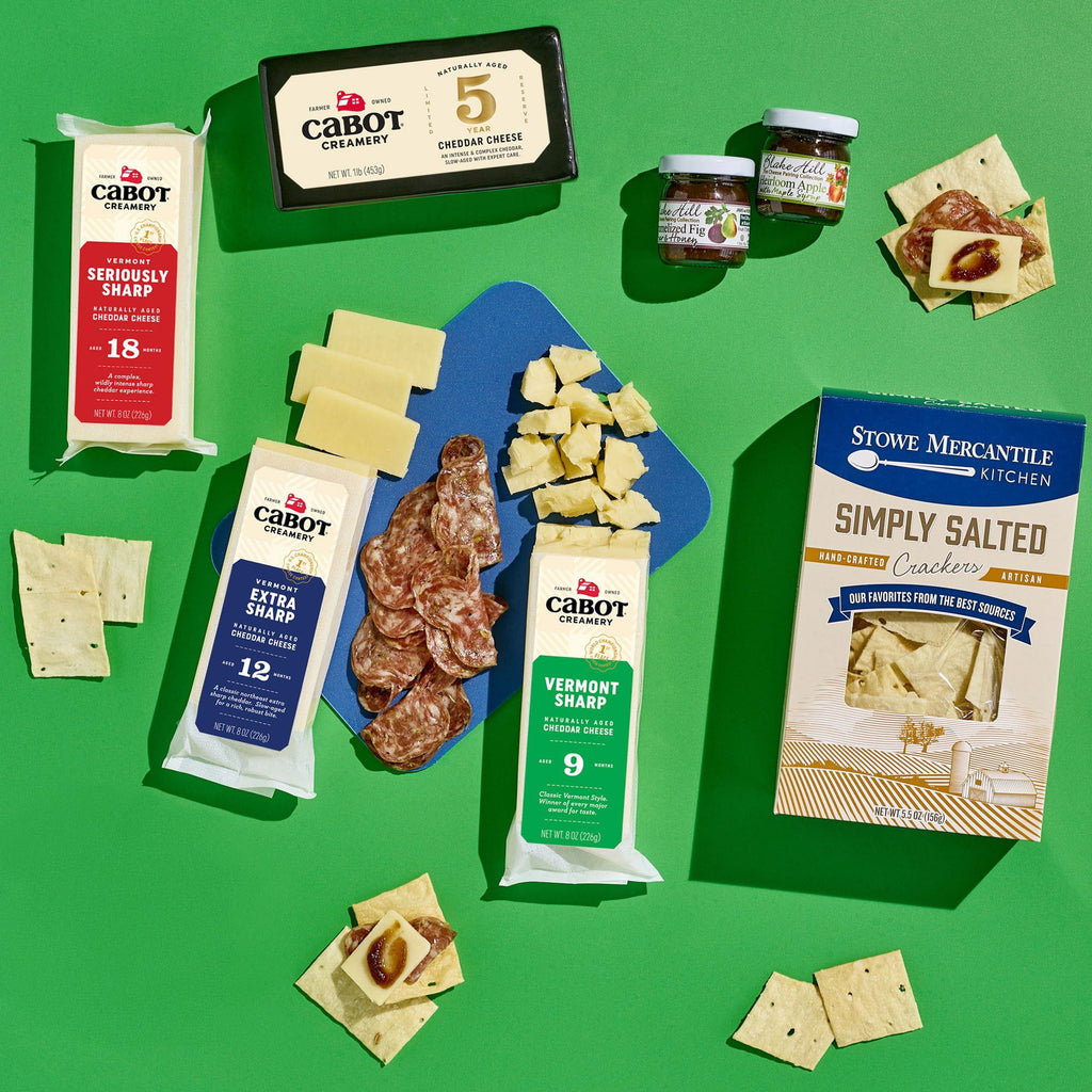 An overhead view of Cabot Creamery’s Classic Vermont Charcuterie Gift Box with cheddar, crackers, salami, jam jars, and Stowe Mercantile Kitchen Simply Salted Crackers on a green background—perfect for a charcuterie basket.