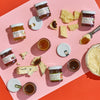The BHP Cabot Pairing Collection by Provisions International features a pink tray displaying assorted cheeses cut into chunks alongside jars of preserves against a red backdrop. The preserves include flavors like fig and raspberry marmalade, with some jars open and lids nearby.