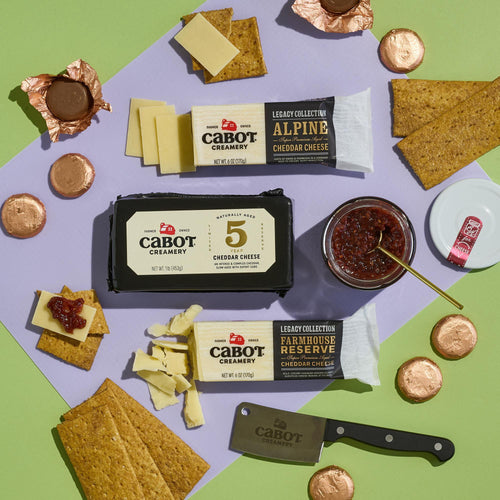 The Salty & Sweet Gift Pack by Cabot Creamery features Vermont cheese, crackers, chocolate coins for pairing, and a jar of jam arranged with a cheese knife and sliced cheese on a pastel surface.