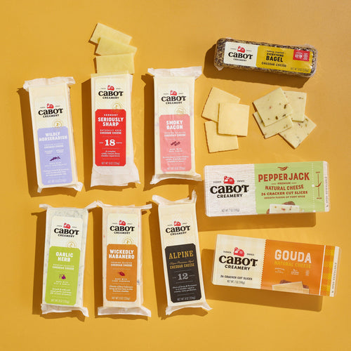 Cabot Creamery’s Fan Favorites Cheese Gift Box is arranged on a yellow surface, showcasing award-winning blocks and slices in flavors like cheddar, Gouda, Smoky Bacon, and Wickedly Habanero alongside crackers.
