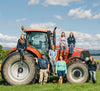 A group of six people pose joyfully with a large red tractor in a grassy field. Four women sit and stand on the tractor, while one man and one woman stand in front. The sky is blue with scattered clouds in the background.