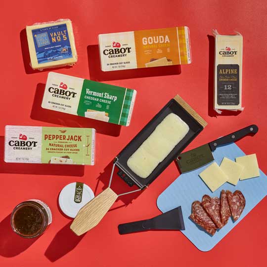 Cabot Cheeses in packaging, with meat, cheese slices, knives, jams and a raclette of melty cheese on a red background.