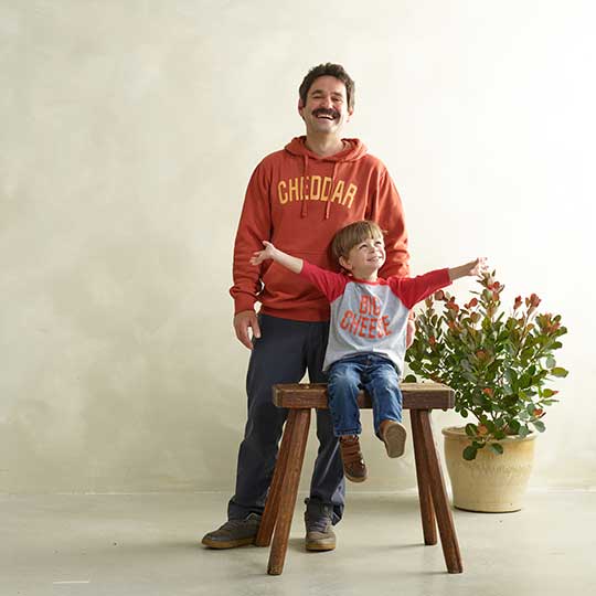 man wearing cheddar hoodie and young boy sitting on a stool wearing a Big Cheese 3/4 Sleeve shirt with a plant next to them.
