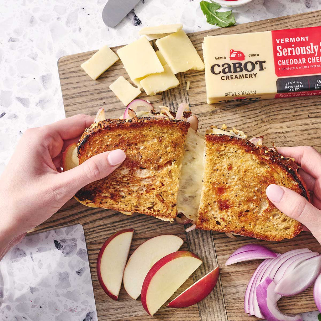 Cabot Seriously Sharp Cheese with a Grilled Cheese sandwich on a cutting board with apples and onions