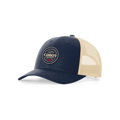 The Committed to Quality Trucker Hat by select design is navy blue and beige with a circular Cabot Creamery patch reading Premium Quality Crafted to Last. It features an adjustable snap back, a mesh rear, and honors Cabot farmers dedication to quality.