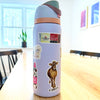 A lavender Owala water bottle is decorated with the Cabot Creamery x Adrianne Sticker Pack by Sticker Giant—featuring cow, butter, cheese, and milk carton designs—on a wooden table in a bright dining room. Ideal for dairy fans.