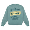 A teal Life’s Better With Butter Crewneck by New Duds, featuring a graphic of a butter stick and the words “Life’s Better With Butter,” “Cabot Creamery,” and “Vermont, USA” on the back, with small branding on one sleeve.