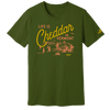 The Life is Cheddar in Vermont T-shirt by New Duds features an olive green color and a yellow-orange barn, trees, and mountains illustration, with the text "Life is Cheddar in Vermont" and Cabot logo below the scene and on the left sleeve.