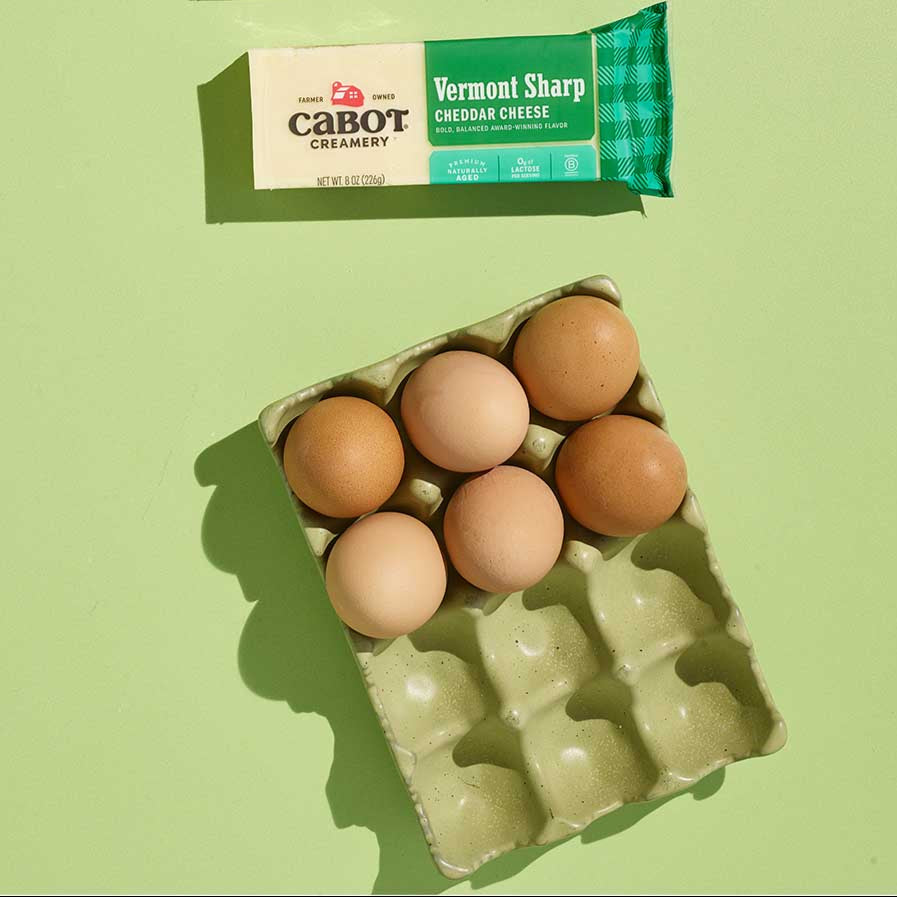 Cabot Lite50 Cheddar Cheese and Cabot Vermont Sharp Cheddar Cheese with eggs in a carton on a green background.