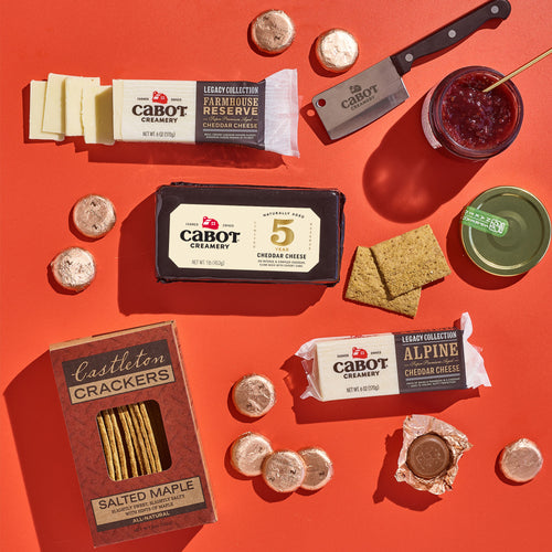 The Salty & Sweet Gift Pack by Cabot Creamery is beautifully arranged on an orange surface, featuring Vermont cheeses such as Cabot cheddar with maple crackers. It includes a jar of Bonne Maman jam, macarons, and a cheese knife, promising indulgence in every bite.