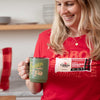 A smiling woman wears a red Seriously Sharp Cheddar Bar T-shirt by New Duds and holds a green mug with Life is Cheddar in Vermont. A Cabot Seriously Sharp Cheddar cheese package is pinned to her shirt, with kitchen shelves and plates behind her.