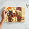 A hand reaches for cheese from the Cabot X That Cheese Plate: Ultimate Charcuterie Gift Box by Cabot Creamery, featuring award-winning cheeses, cured meats, crackers, dried fruit, olives, nuts, and dip on a white marble surface.