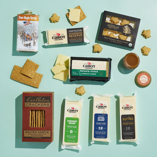 A flat lay of the Cabot Creamery Vermont Maple & Cheese Gift Box, featuring cheddar cheese, maple syrup, salted maple crackers, and Vermont maple candy on a light blue background with scattered cheese and cracker pieces.