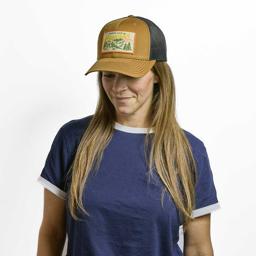 A person wearing a New Duds Where Life is Cheddar Trucker Hat featuring a brown and black mesh back and a mountain landscape patch.