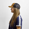 Side profile of a person wearing a brown and dark blue Where Life is Cheddar Trucker Hat with a patch on the front and text embroidered on the side.