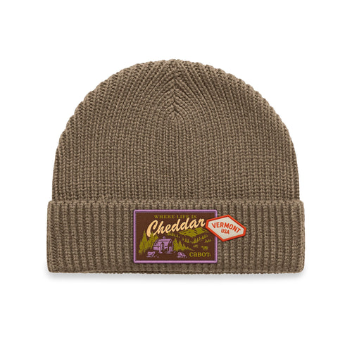 The Where Life is Cheddar Beanie by New Duds is a brown knit beanie with a folded brim, featuring two patches: a purple and yellow patch reading “Cheddar,” and a red, Vermont-shaped patch labeled “Vermont USA.”.