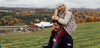 A man carries a young girl on his shoulders in a field during autumn. The girl wears a white beanie and patterned dress. The background features hills with colorful fall foliage and a barn, under a cloudy sky.