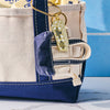 A close-up of a canvas tote with navy trim holds Cabot butter sticks. A WizardPins Butter Charm Keychain, shaped like a butter stick, hangs from the handle. The blue background and cracked white surface complete the scene.