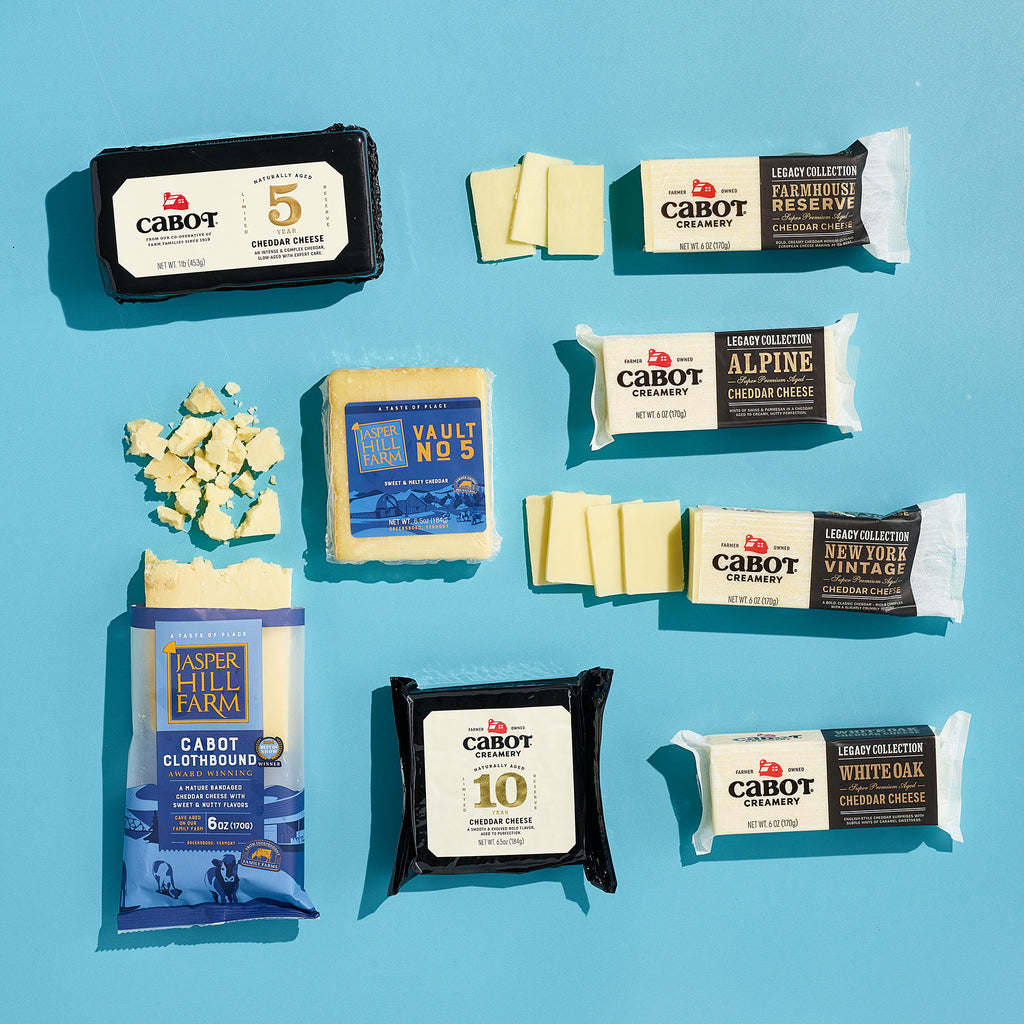 Assorted Cheese, Please Cheddar Medley blocks from Cabot Creamery are beautifully packaged on a blue background. Includes naturally aged cheddar and parmesan varieties, with some blocks unwrapped to reveal slices and crumbles.